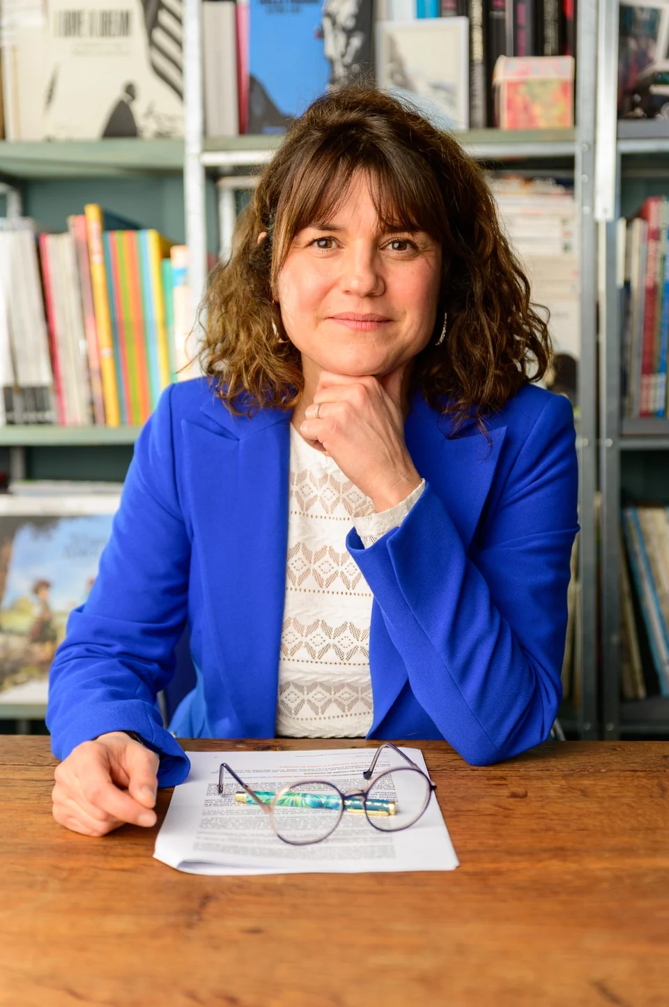 Julia Peironet-Brémond en veste bleue, la main sous le menton devant sa bibliothèque
