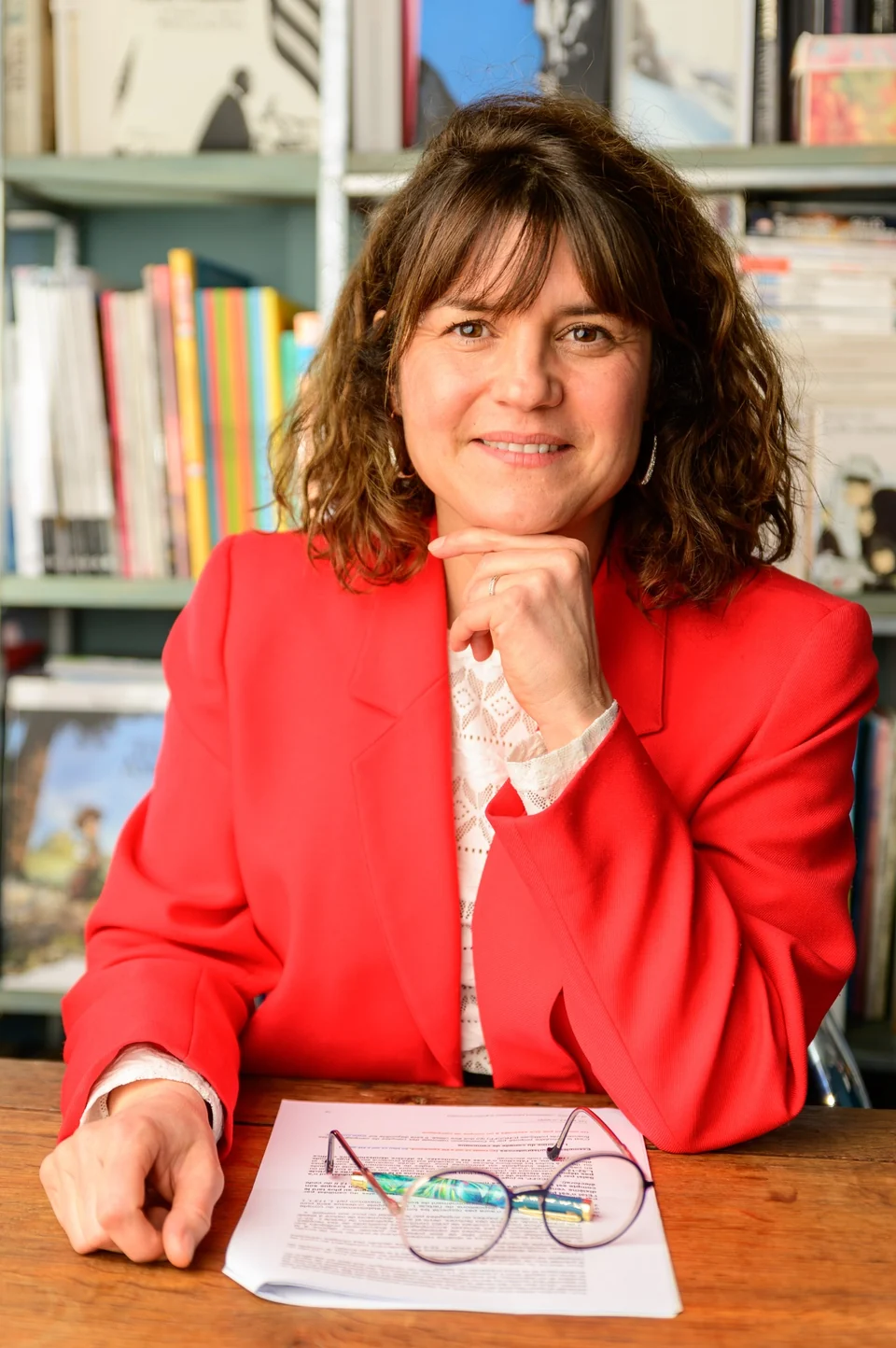 Julia Peironet-Brémond en veste rouge, la main sous le menton, devant sa bibliothèque